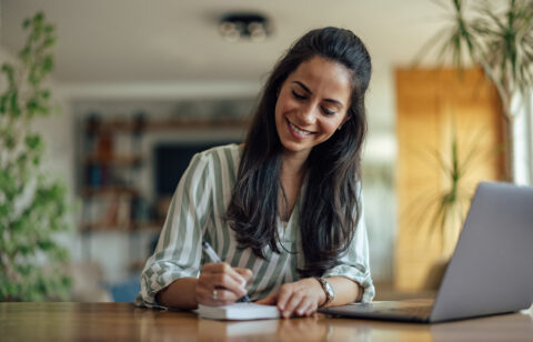 Woman smiling while writing in a notebook at a table with a laptop nearby.