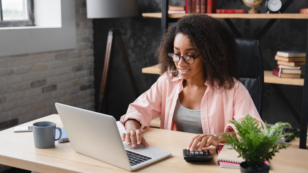 Woman working at a desk with a laptop and calculator, managing finances in a home office.