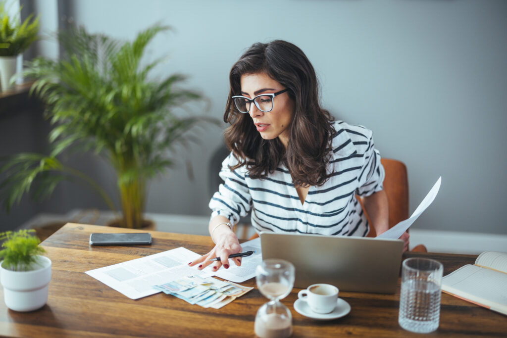 Woman reviewing paperwork and cash at a table while using a laptop.