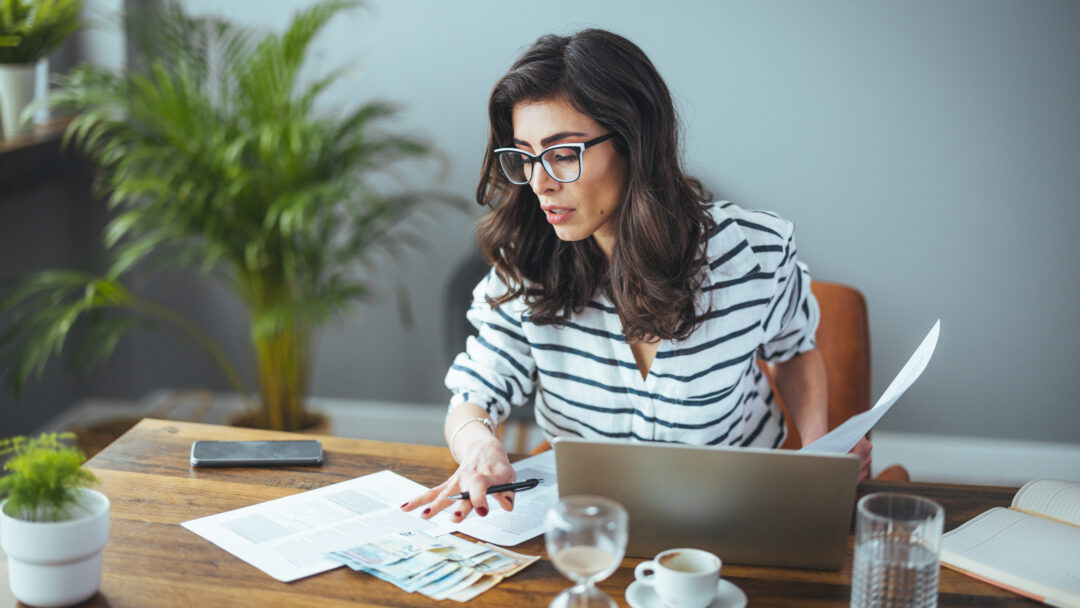 Woman reviewing paperwork and cash at a table while using a laptop.