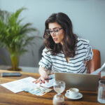 Woman reviewing paperwork and cash at a table while using a laptop.