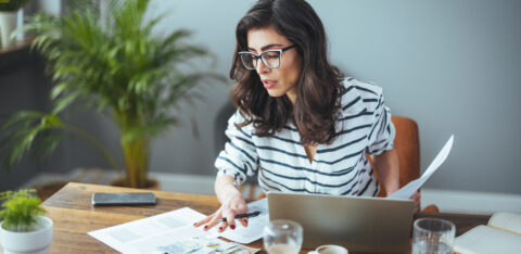Woman reviewing paperwork and cash at a table while using a laptop.
