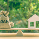 Small sack labeled “Loan” balanced on a scale opposite a model house, representing borrowing for a home.