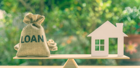 Small sack labeled “Loan” balanced on a scale opposite a model house, representing borrowing for a home.