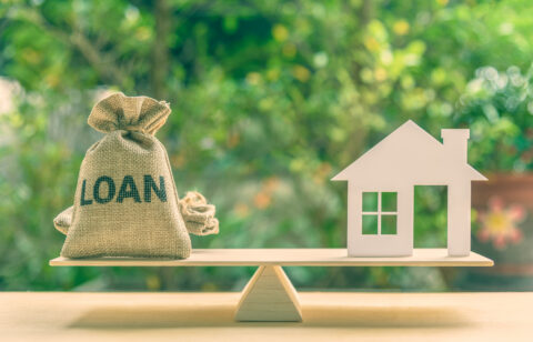 Small sack labeled “Loan” balanced on a scale opposite a model house, representing borrowing for a home.