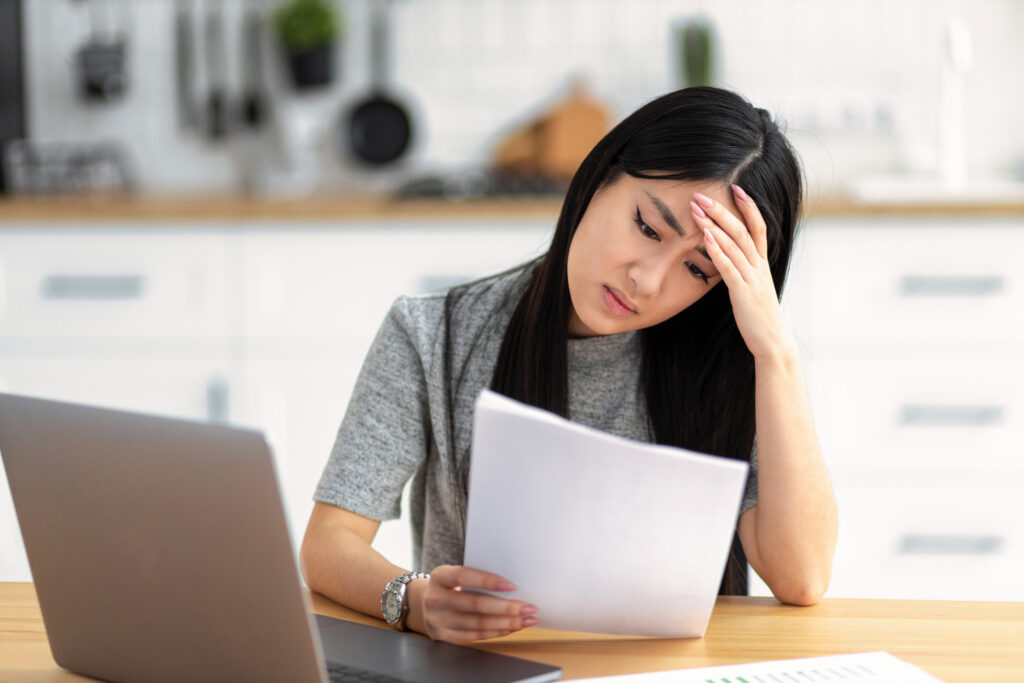 Woman looking stressed while reading a document at a table with a laptop nearby.