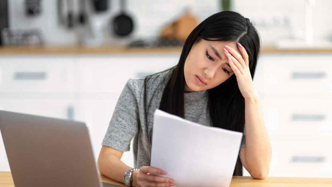 Woman looking stressed while reading a document at a table with a laptop nearby.