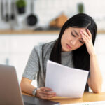 Woman looking stressed while reading a document at a table with a laptop nearby.