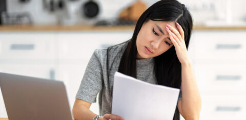 Woman looking stressed while reading a document at a table with a laptop nearby.