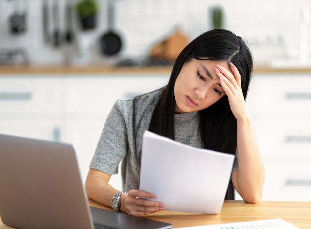 Woman looking stressed while reading a document at a table with a laptop nearby.