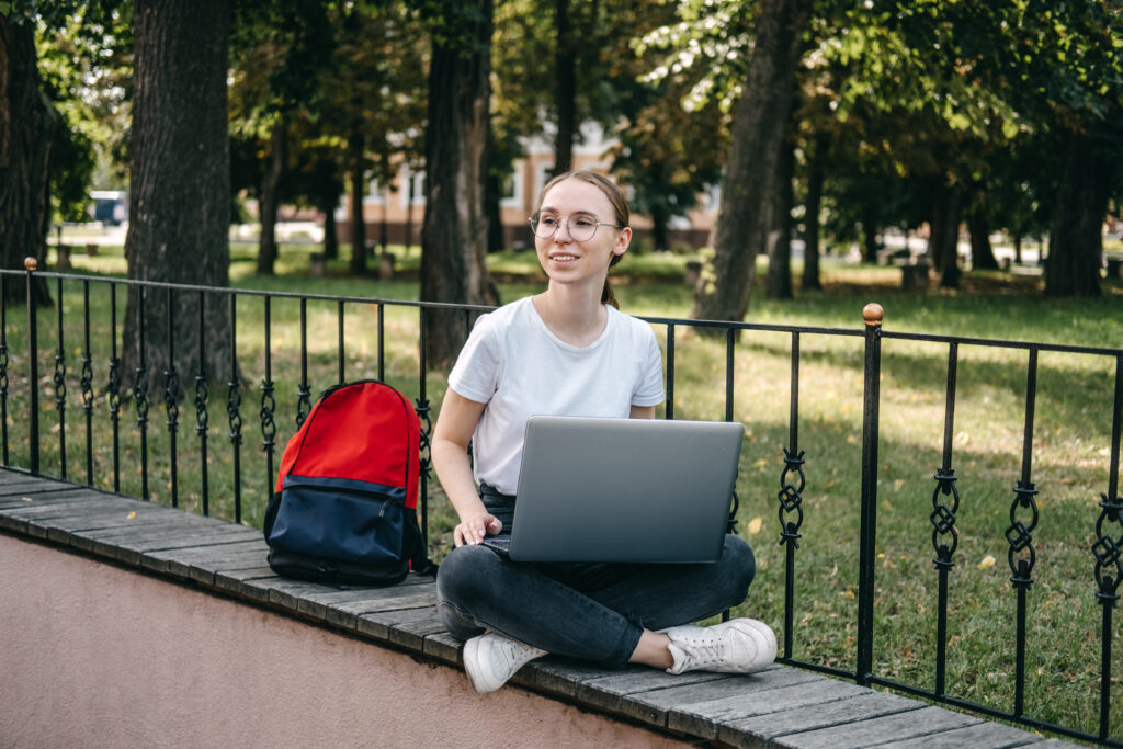 Student sitting outdoors with a laptop on a ledge, with a backpack beside her.