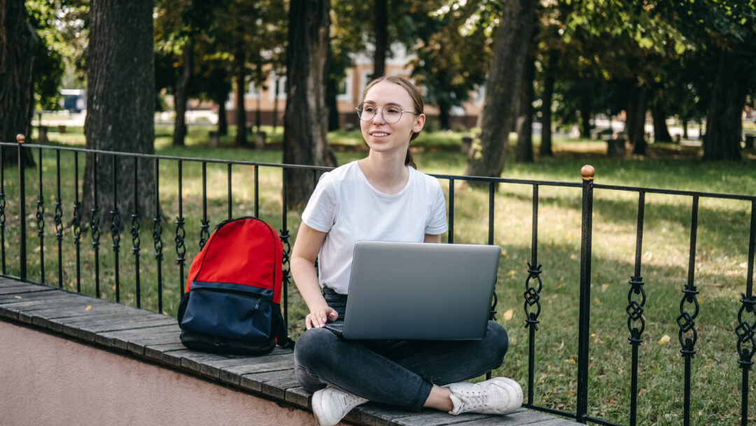 Student sitting outdoors with a laptop on a ledge, with a backpack beside her.