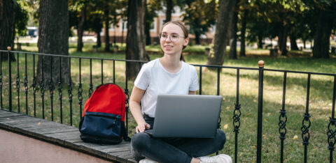 Student sitting outdoors with a laptop on a ledge, with a backpack beside her.
