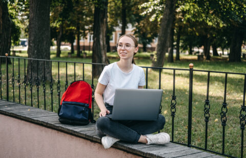 Student sitting outdoors with a laptop on a ledge, with a backpack beside her.