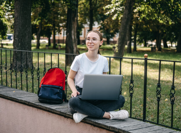 Student sitting outdoors with a laptop on a ledge, with a backpack beside her.