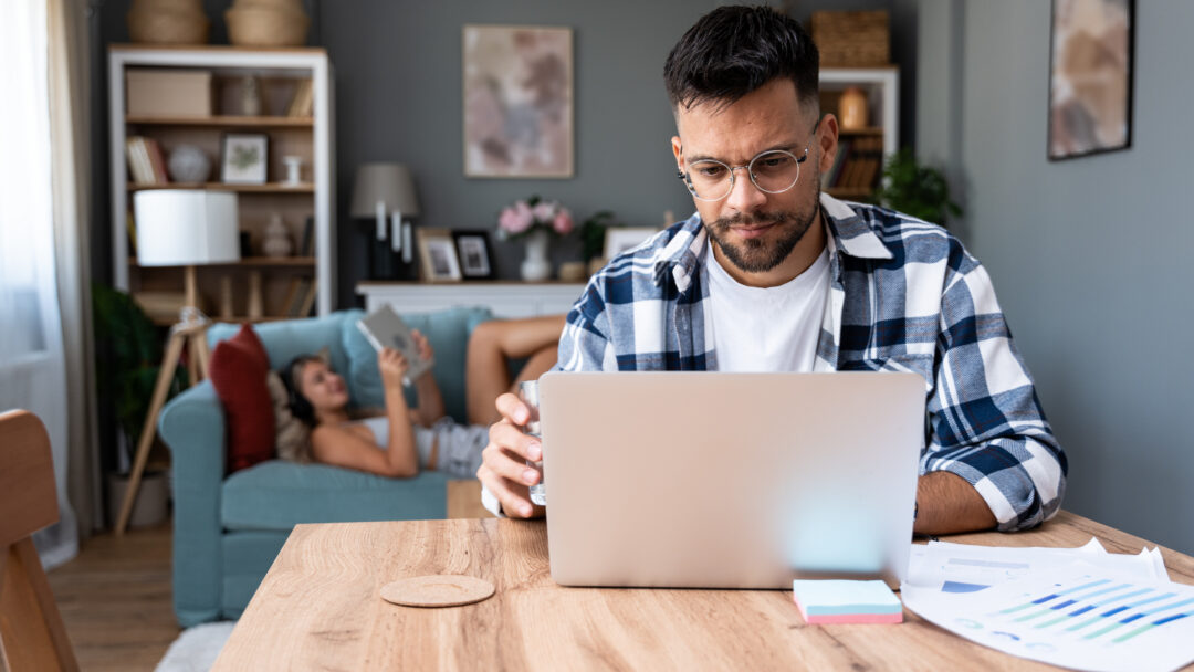 Man working on a laptop at a table while holding a glass of water, with a woman relaxing on a couch in the background.