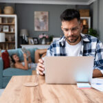 Man working on a laptop at a table while holding a glass of water, with a woman relaxing on a couch in the background.