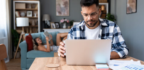 Man working on a laptop at a table while holding a glass of water, with a woman relaxing on a couch in the background.