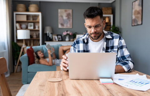 Man working on a laptop at a table while holding a glass of water, with a woman relaxing on a couch in the background.