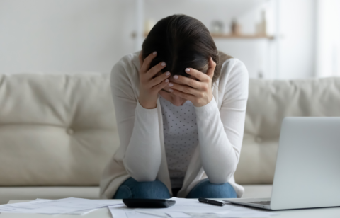Person sitting on a couch with head in hands, surrounded by bills, a calculator, and a laptop.