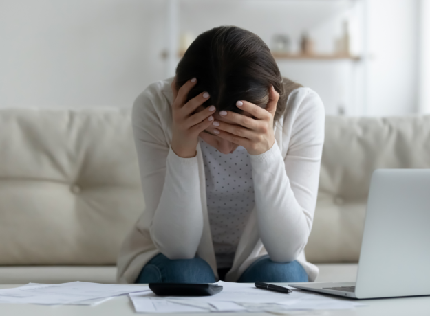 Person sitting on a couch with head in hands, surrounded by bills, a calculator, and a laptop.