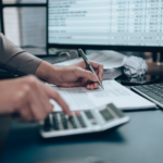 Person using a calculator and writing on paperwork at a desk with financial data on a computer screen