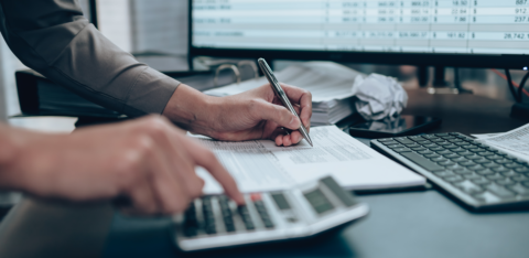 Person using a calculator and writing on paperwork at a desk with financial data on a computer screen
