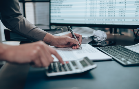 Person using a calculator and writing on paperwork at a desk with financial data on a computer screen