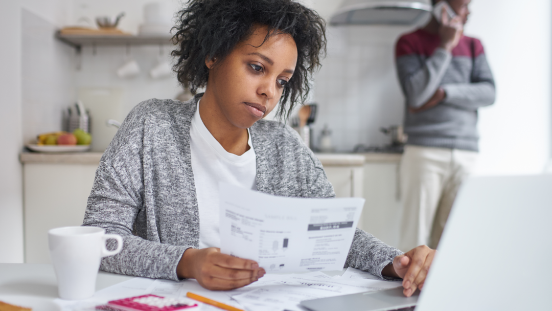 Woman reviewing bills at a table with a laptop while another person stands in the background on the phone.