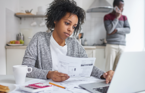 Woman reviewing bills at a table with a laptop while another person stands in the background on the phone.