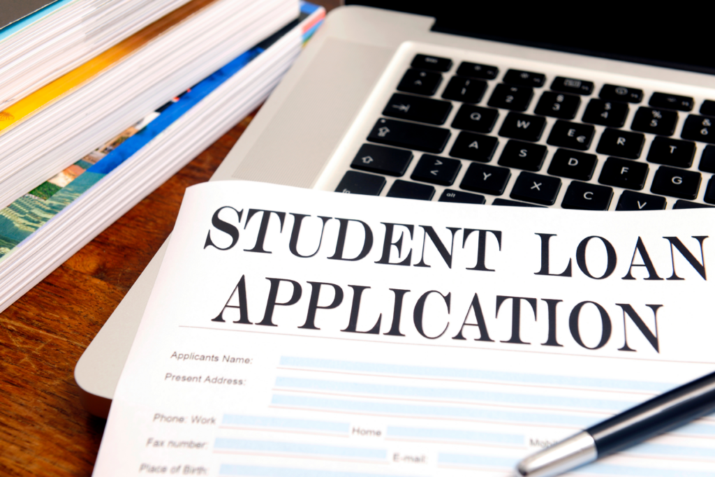 Student loan application form on a desk next to a laptop, pen, and stacked books