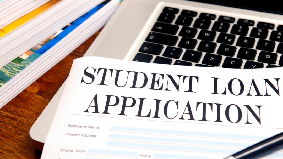 Student loan application form on a desk next to a laptop, pen, and stacked books