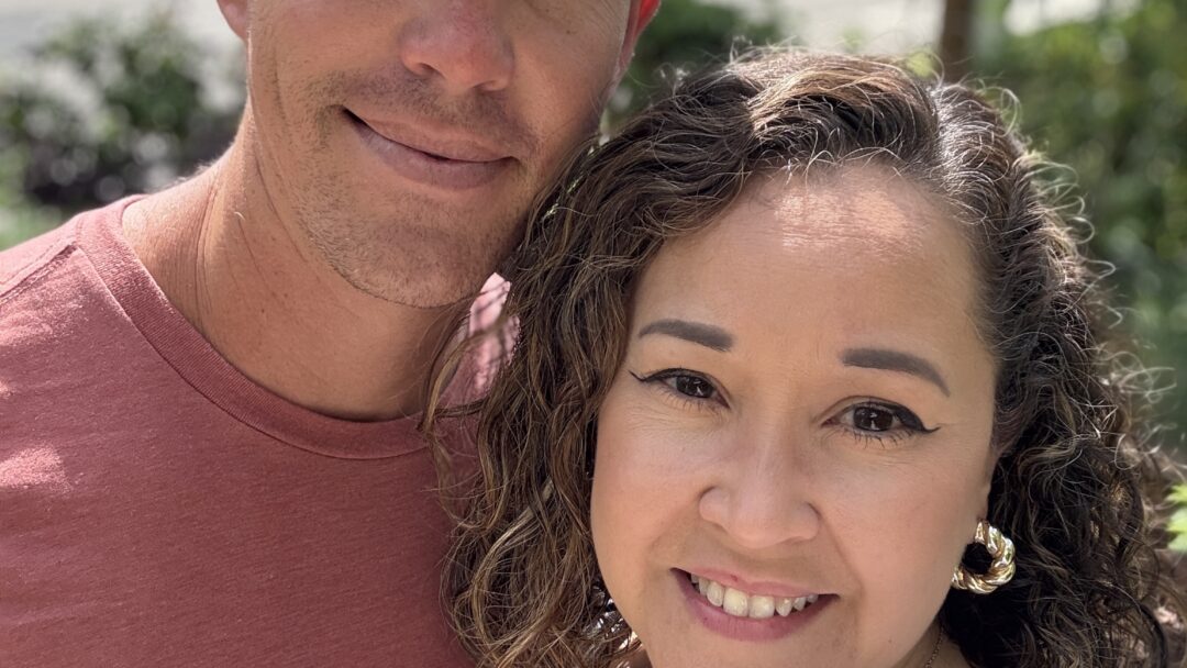Smiling couple taking a close-up selfie outdoors with trees and sunlight in the background