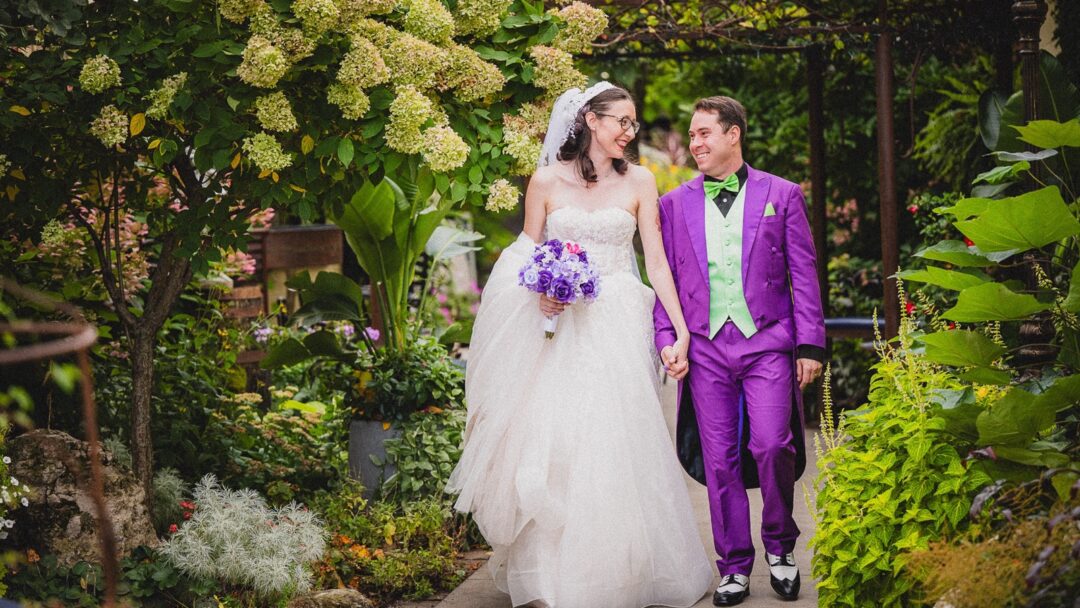 Bride and groom walking hand in hand through a garden, smiling at each other.
