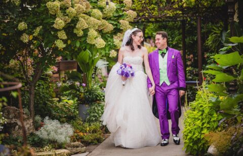 Bride and groom walking hand in hand through a garden, smiling at each other.