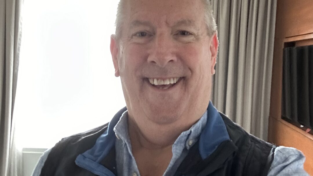 Smiling man taking a selfie indoors with a window and curtains in the background.