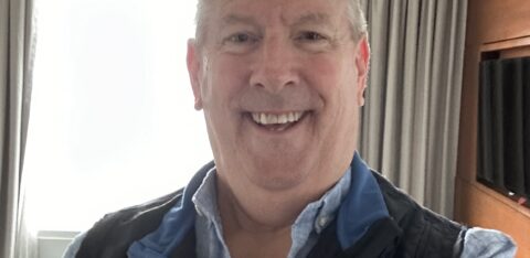 Smiling man taking a selfie indoors with a window and curtains in the background.