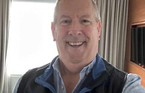 Smiling man taking a selfie indoors with a window and curtains in the background.