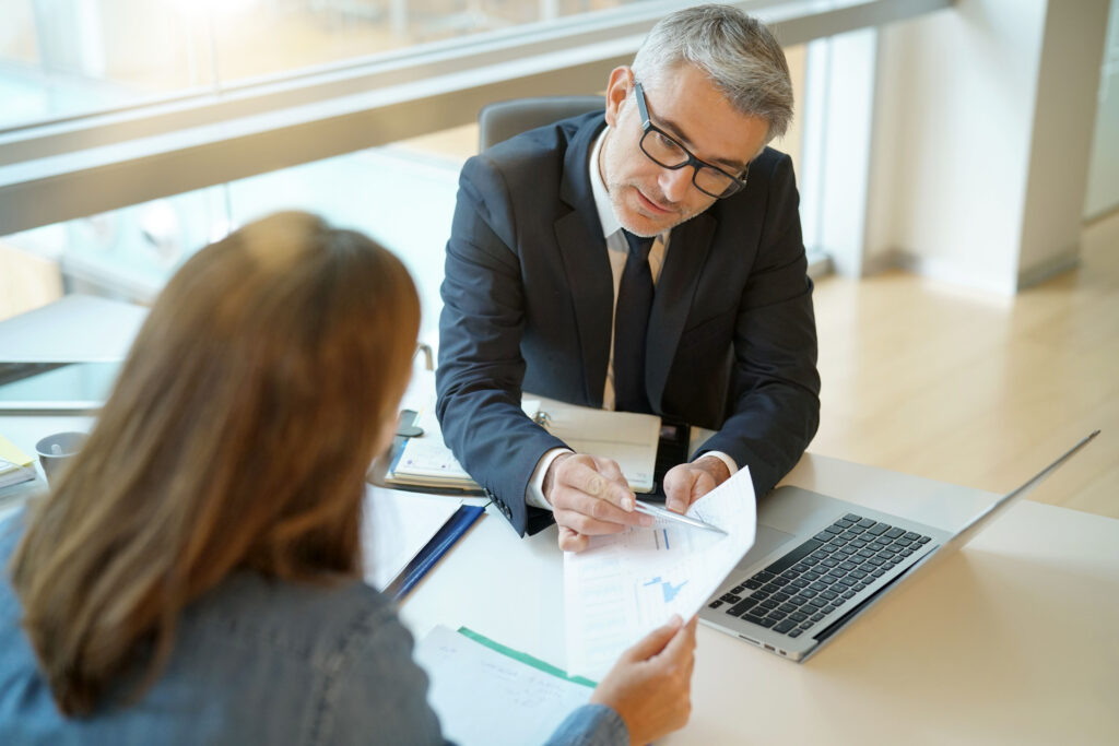 Man in a suit reviewing documents with a client at a desk, pointing to a chart on the paper.
