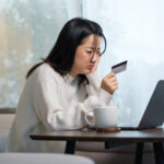 Person sitting at a table using a laptop while holding a credit card, appearing focused