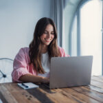 Person smiling while using a laptop at a wooden table, with a notebook nearby