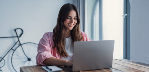 Person smiling while using a laptop at a wooden table, with a notebook nearby