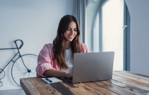 Person smiling while using a laptop at a wooden table, with a notebook nearby