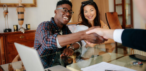 Couple smiling while shaking hands with a professional across a table with paperwork and a laptop