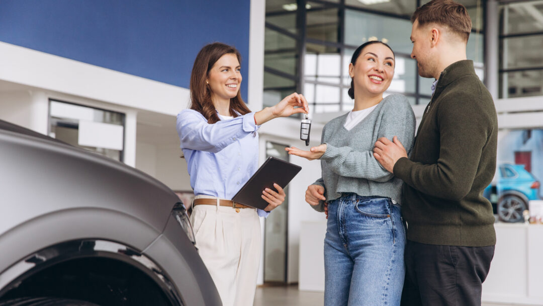 Saleswoman handing Salesperson handing car keys to a smiling couple at a dealership, with a vehicle nearby.