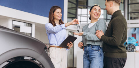 Saleswoman handing Salesperson handing car keys to a smiling couple at a dealership, with a vehicle nearby.