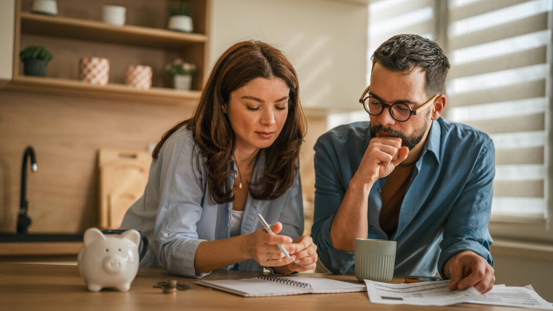 Two adults looking over financial documents and taking notes at home, with coins and a piggy bank on the table.