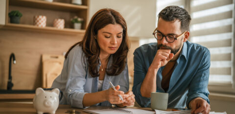 Two adults looking over financial documents and taking notes at home, with coins and a piggy bank on the table.