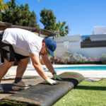 Middle aged man removing artificial grass in a sunny backyard with a swimming pool, showing residential landscaping work, lawn replacement and outdoor home improvement project