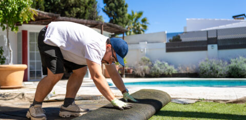 Middle aged man removing artificial grass in a sunny backyard with a swimming pool, showing residential landscaping work, lawn replacement and outdoor home improvement project